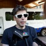 Silhouette of Jony Ive examining a conceptual screenless AI device on a desk with abstract branding background