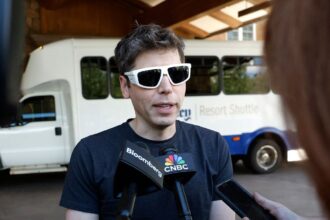 Silhouette of Jony Ive examining a conceptual screenless AI device on a desk with abstract branding background