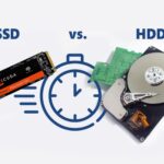 A close-up of a high-speed NVMe SSD next to a blurred gaming monitor showing the Grand Theft Auto V loading screen, illustrating the struggle of game storage.