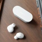 A pair of Sony WF-1000XM6 wireless earbuds sitting next to the dual-screen ASUS Zenbook Duo laptop on a minimalist coffee shop table.