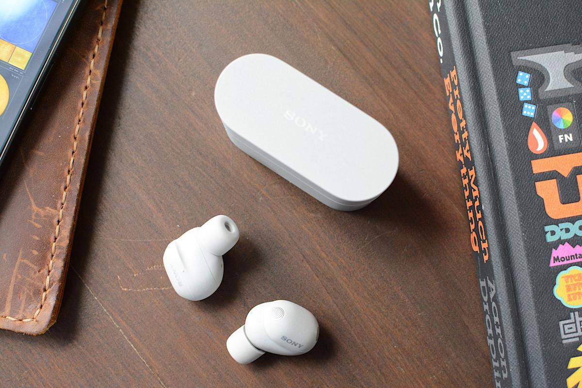 A pair of Sony WF-1000XM6 wireless earbuds sitting next to the dual-screen ASUS Zenbook Duo laptop on a minimalist coffee shop table.