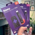 A person holding a slim Roku streaming stick next to an older television set